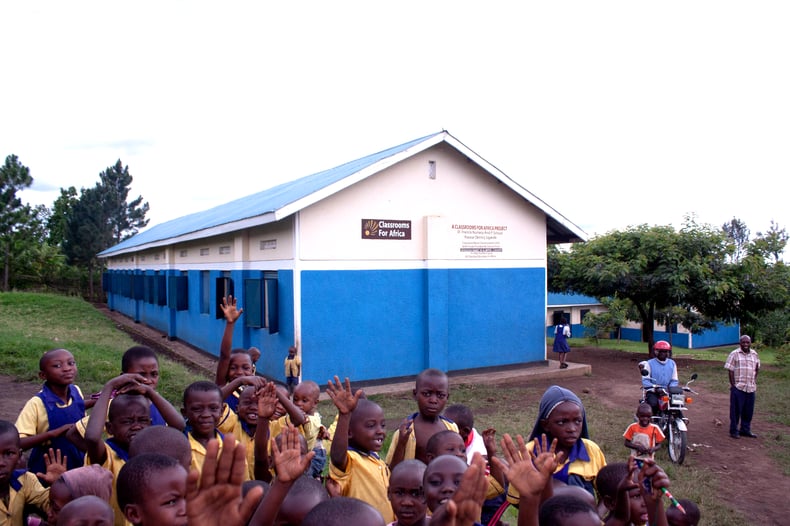 A group of excited children gather in front of a Classrooms for Africa school building. The children wave happily at the camera.